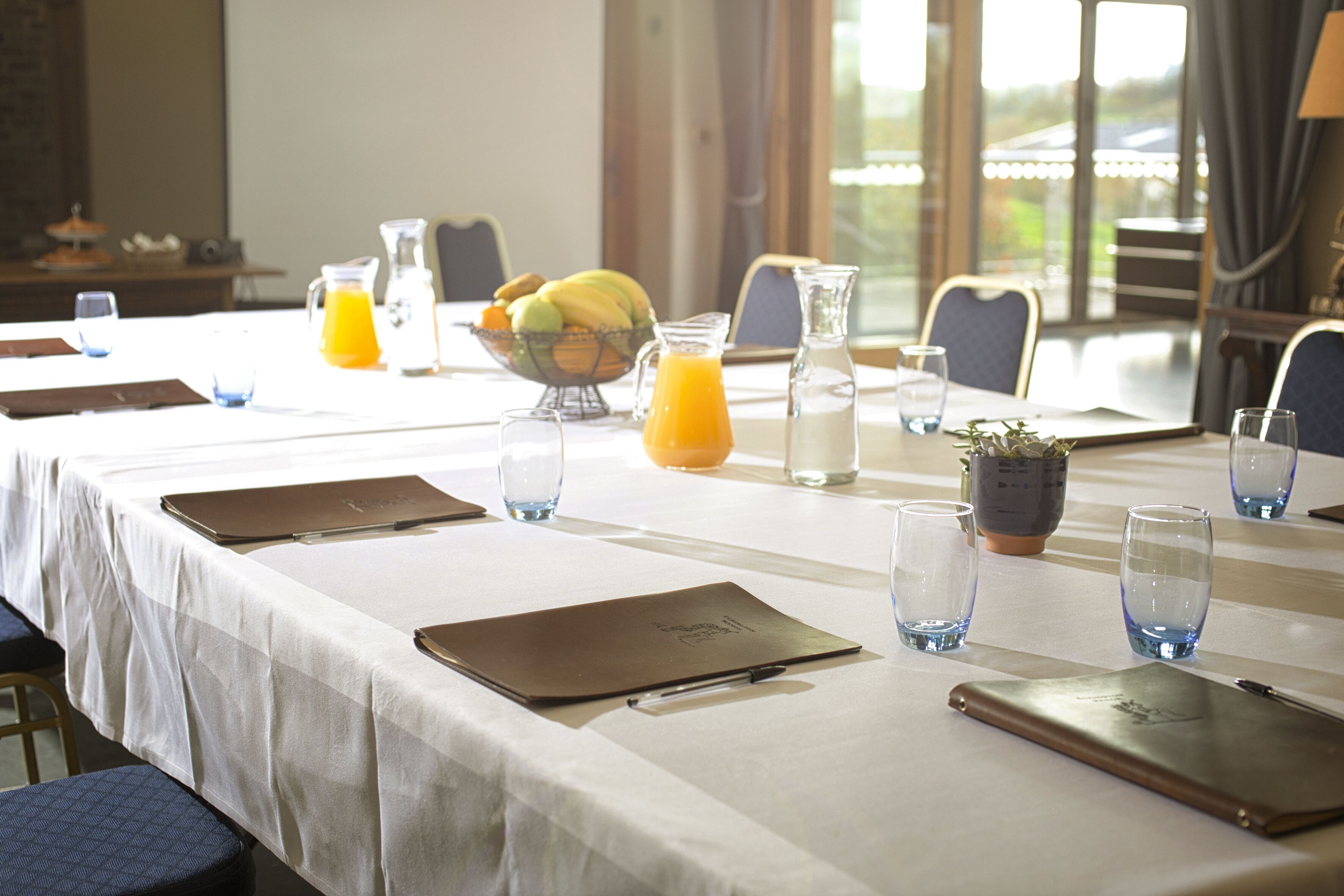 Un salón de conferencias con una larga mesa dispuesta para una reunión, equipado con carpetas, cuadernos y una jarra de jugo de naranja.