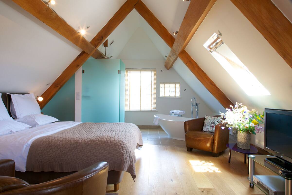 A bedroom with a bed, a chair, a TV, a bathroom with a bathtub, and a window.