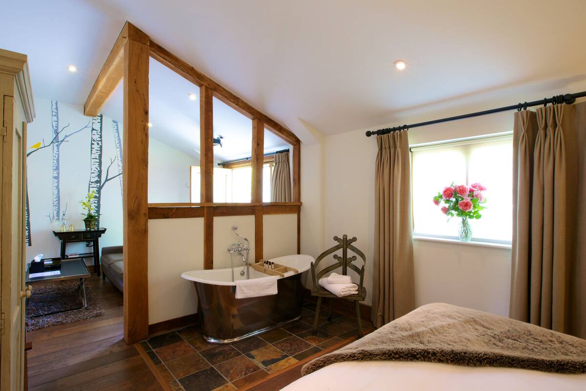 A bathroom with a freestanding bathtub, a wooden chair, a vanity with a mirror, and a window with curtains.
