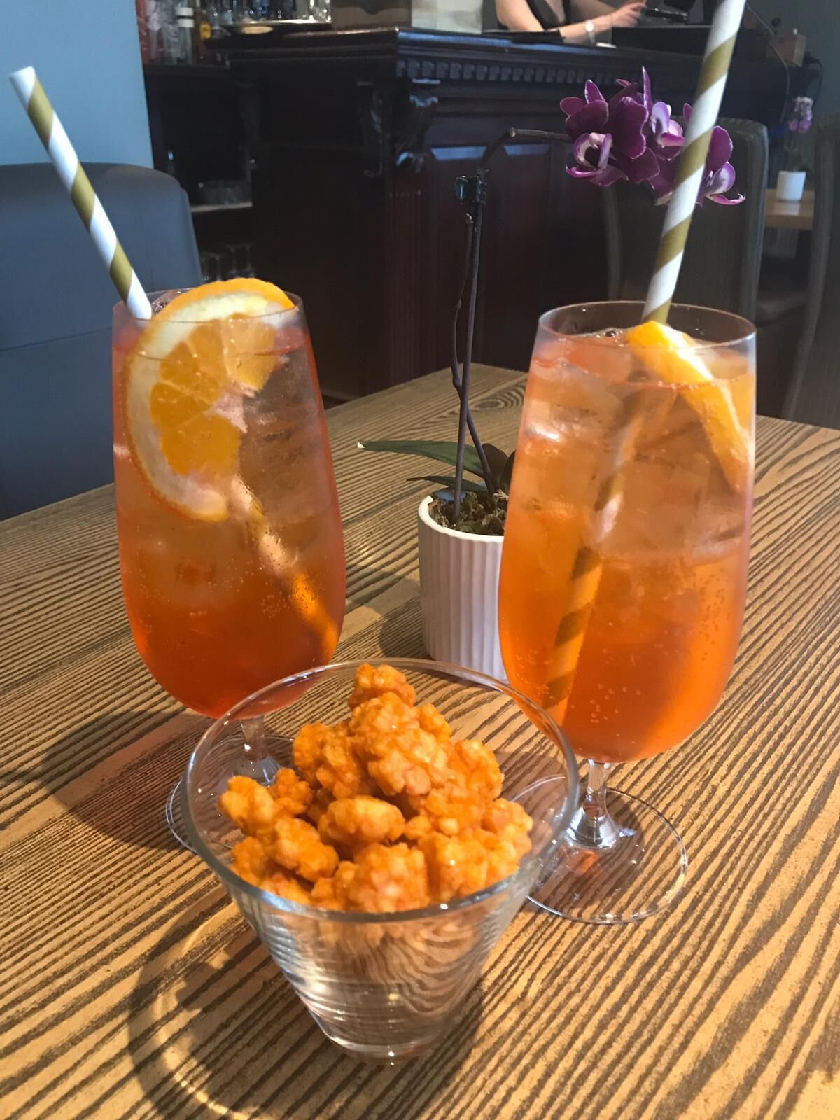 Two drinks with orange slices and straws, a bowl of fritters, and a bar area in the background.
