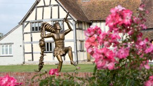 Una estatua de bronce de una figura musculosa con una lanza, ubicada frente a un edificio de estilo Tudor con una chimenea de ladrillo.