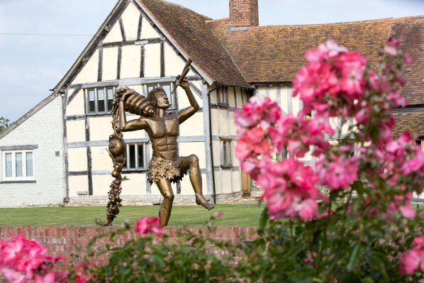 A bronze statue of a muscular figure with a spear, positioned in front of a Tudor-style building with a brick chimney. - Eckington Manor (Pershore)