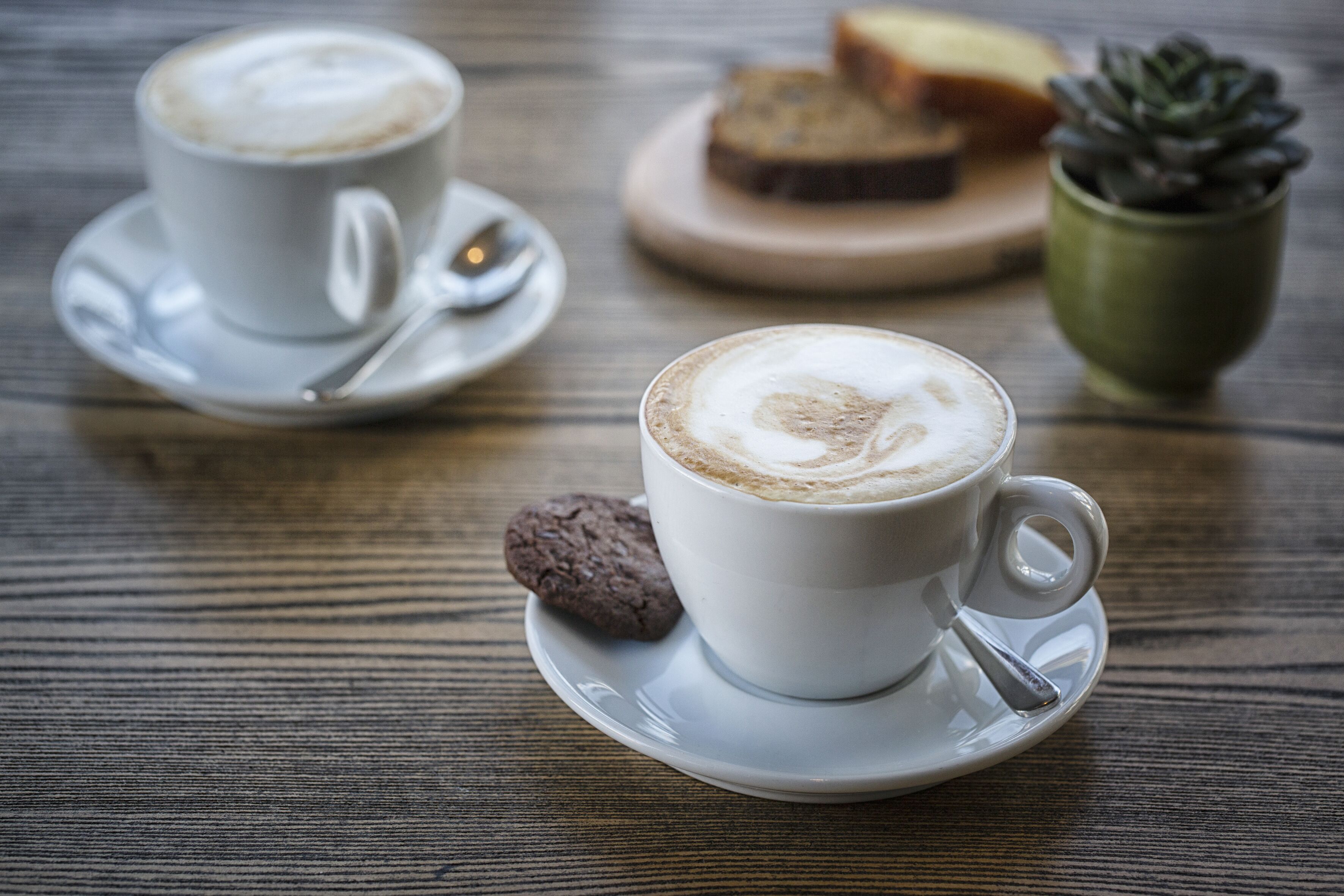 Deux tasses de café blanches, accompagnées de leurs soucoupes et de cuillères, sont posées sur une table en bois.
