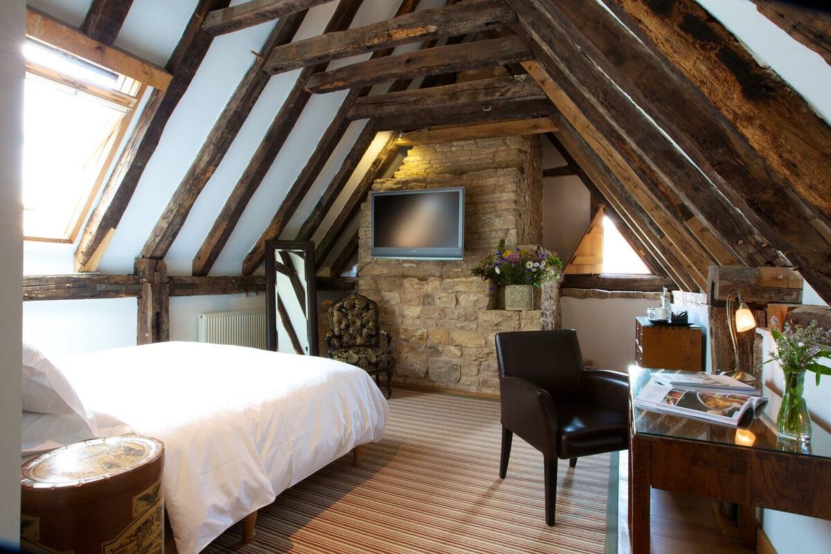 A cozy attic bedroom with a bed, a chair, a desk with a lamp, a TV, and a stone wall.