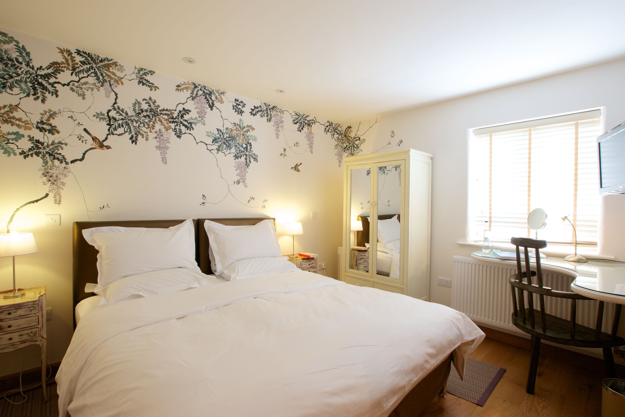 A neatly made bed with white linens, a wooden nightstand with a lamp, a mirror, and a chair in a room with a radiator and a window with blinds.
