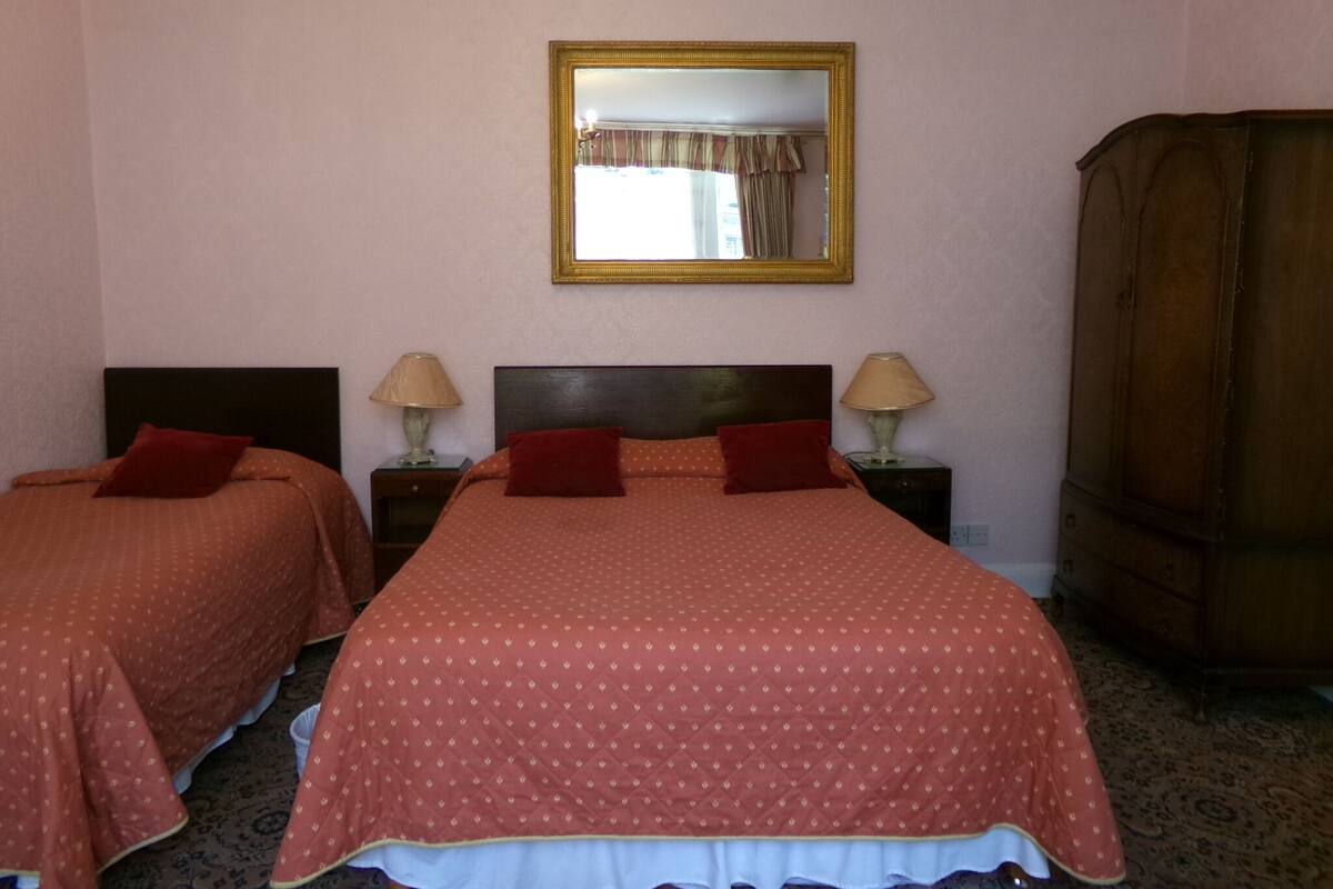 Habitación de hotel con dos camas, cada una con colcha y almohadas rojas a juego, flanqueadas por mesitas de noche con lámparas. Un espejo grande cuelga sobre las camas, y un armario de madera se encuentra a la derecha.