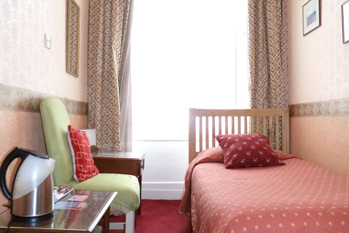 Un dormitorio con cama, una silla, una mesa con hervidor y cortinas.