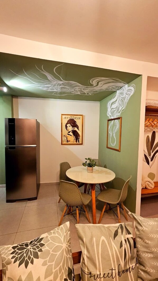 Un espacio habitable compacto con zona de comedor que incluye una mesa redonda y cuatro sillas, refrigerador de acero inoxidable integrado y una pared decorada con un cuadro abstracto de gran tamaño.