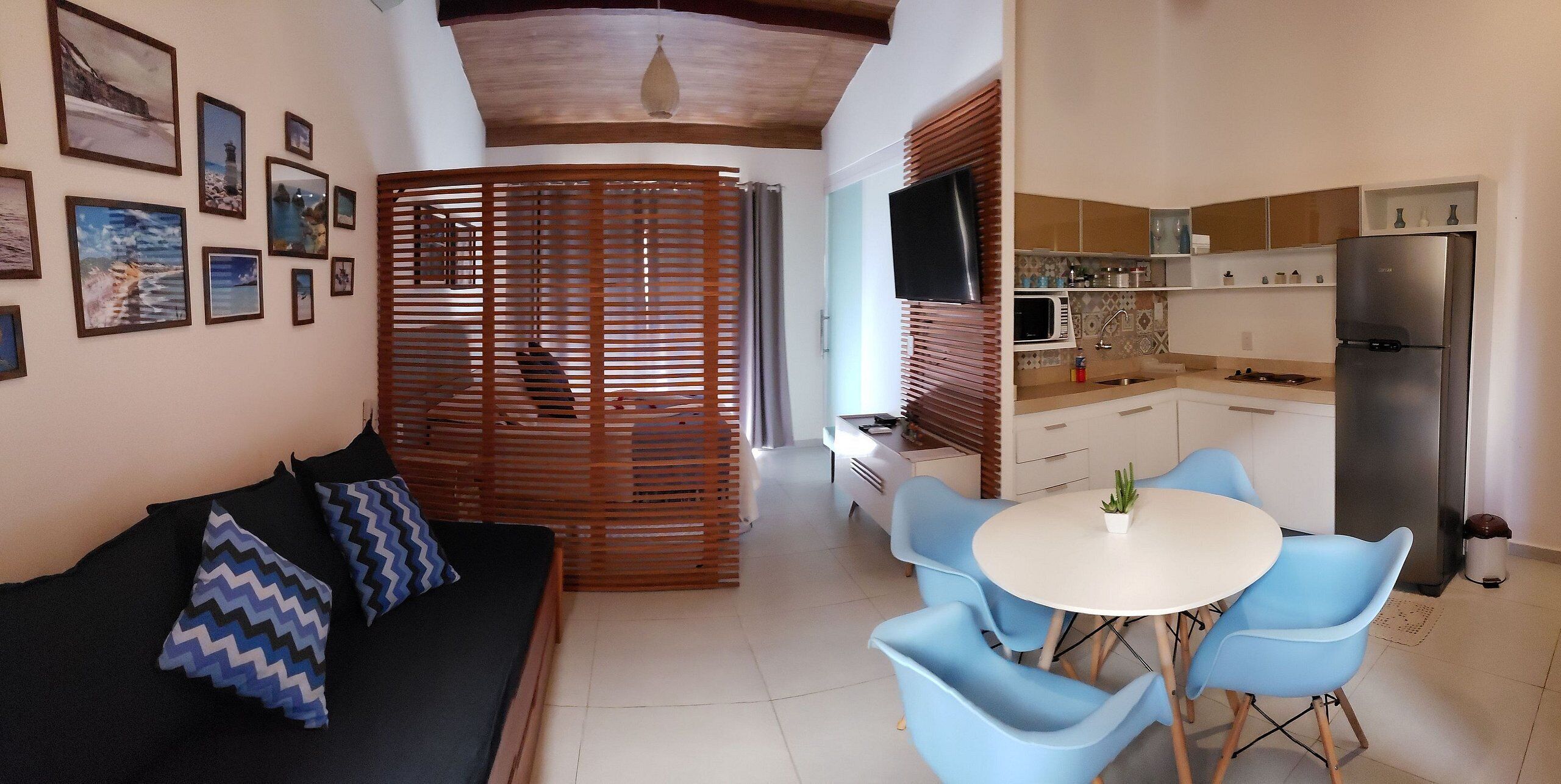Een moderne woonkamer met een bank, blauwe stoelen en een eettafel. In de achtergrond is een keuken met een koelkast en magnetron zichtbaar.