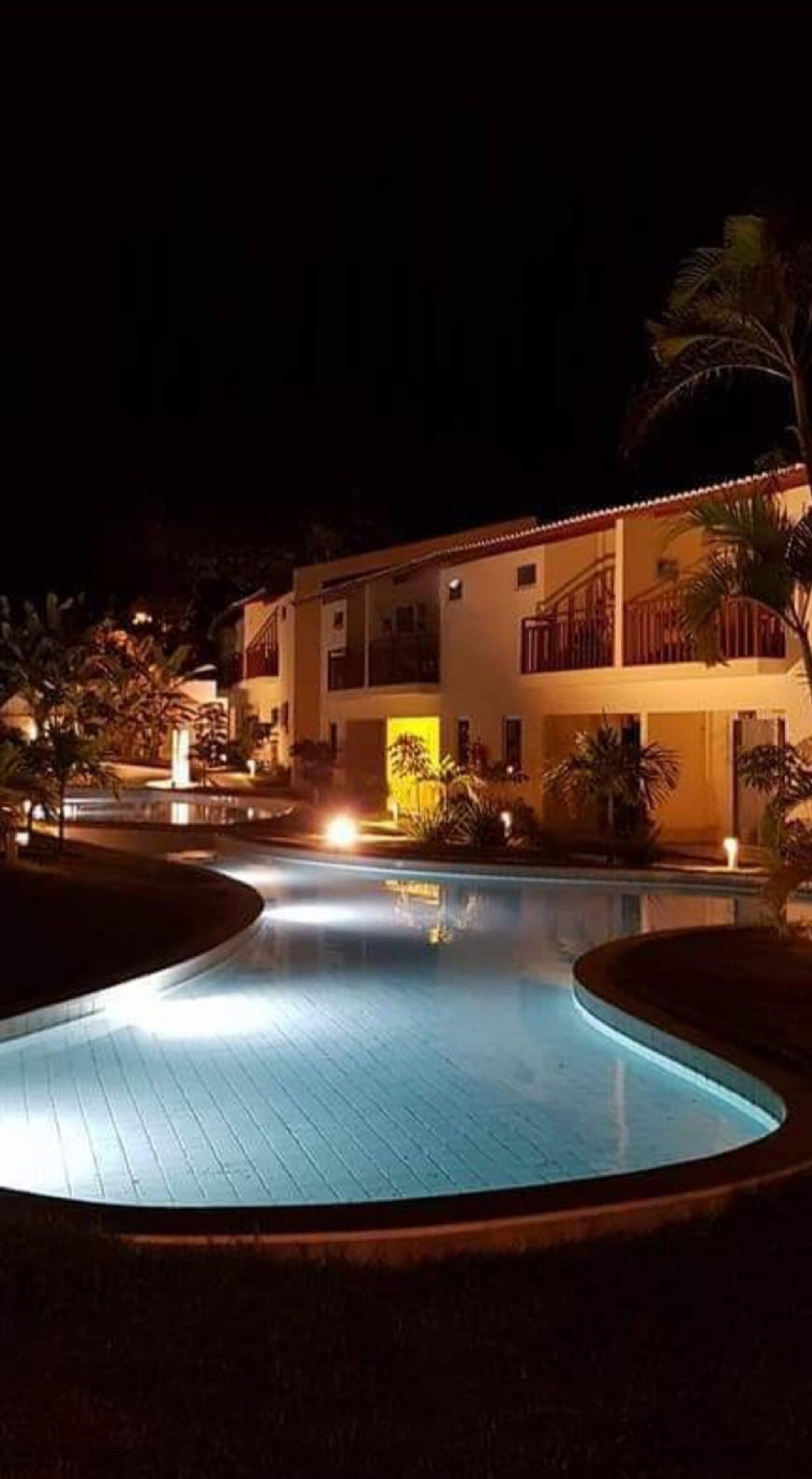 Vista nocturna de un complejo turístico con piscina, un edificio de dos pisos y palmeras.