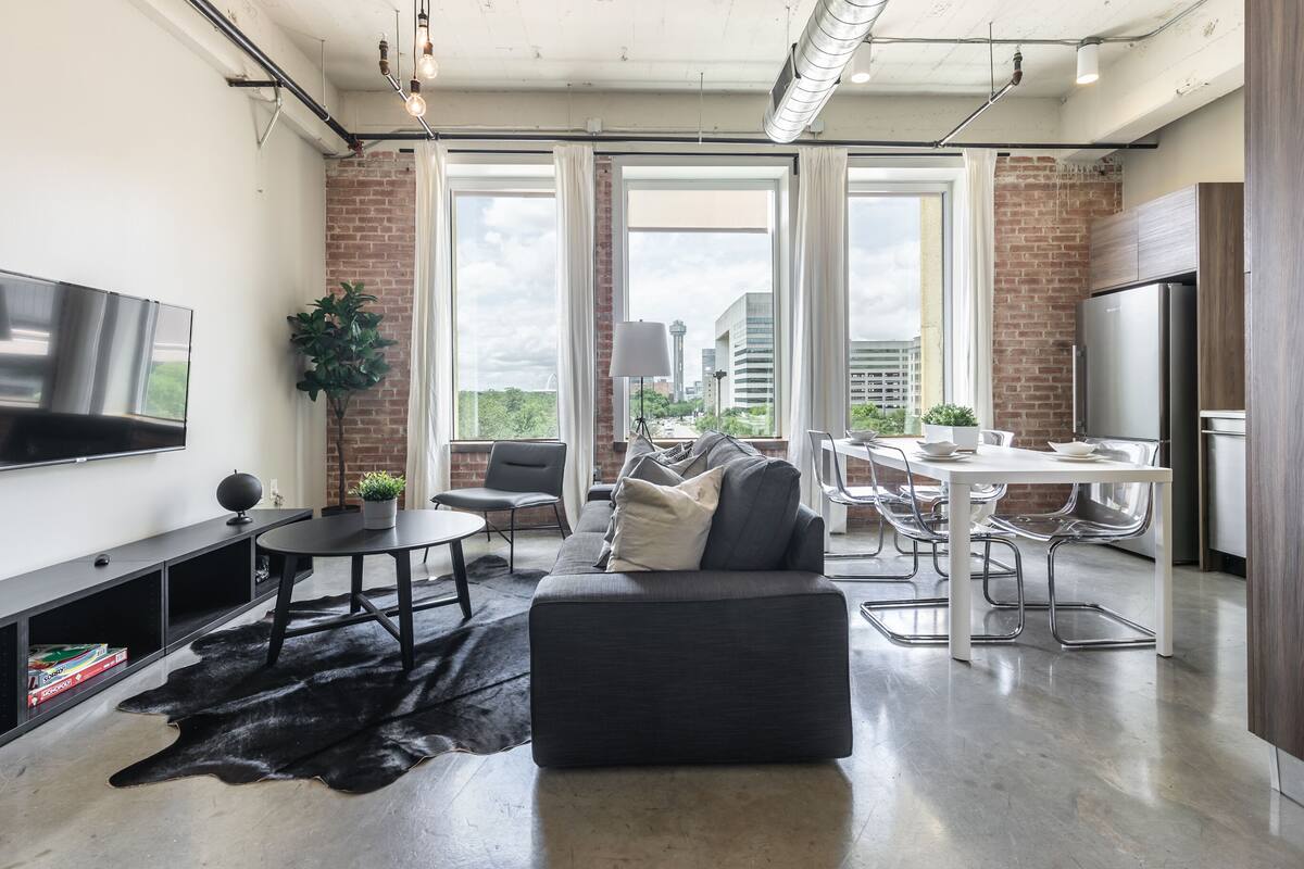 A modern living room with a sofa, dining table, and chairs, large windows with a city view, a flat-screen TV, and a refrigerator.