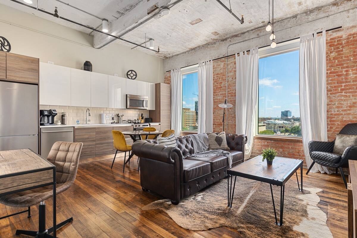 A modern kitchen with stainless steel appliances, a dining area with a wooden table and chairs, a leather sofa, and a large window with a city view.