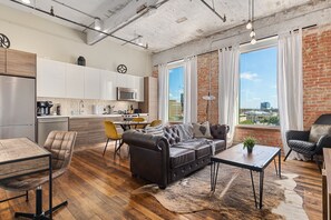 A modern kitchen with stainless steel appliances, a dining area with a wooden table and chairs, a leather sofa, and a large window with a city view. - Downtown Luxury Lofts By RBA Living (Dallas)