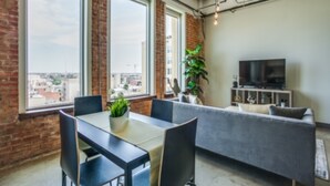 Una sala de estar moderna con una mesa de comedor, sillas, un sofá gris, un televisor y ventanas amplias con vistas a la ciudad.