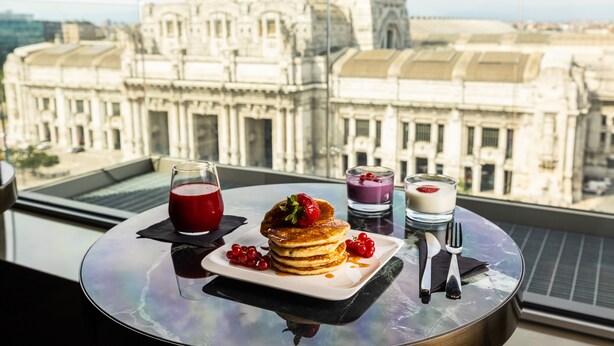 Une table dressée pour le petit-déjeuner, avec des crêpes, des fruits rouges et des boissons, et une vue sur la ville.