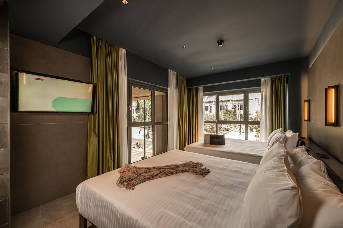 A hotel room with two beds, a flat-screen TV mounted on the wall, and a window with curtains.