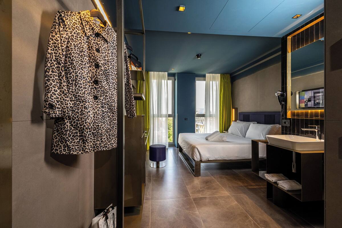 A modern hotel room with a bed, a vanity with a mirror, and a view of the city through large windows.