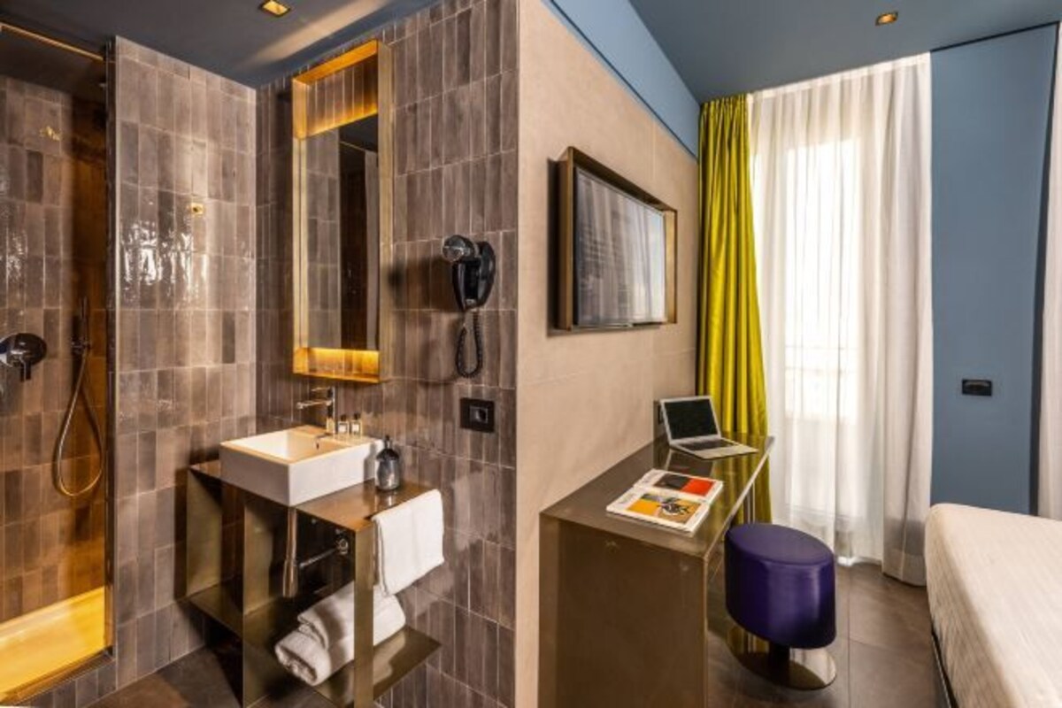 A modern bathroom with a large mirror, a sink, and a shower. There is a small desk with a laptop and a magazine, and a bed with a blue headboard.