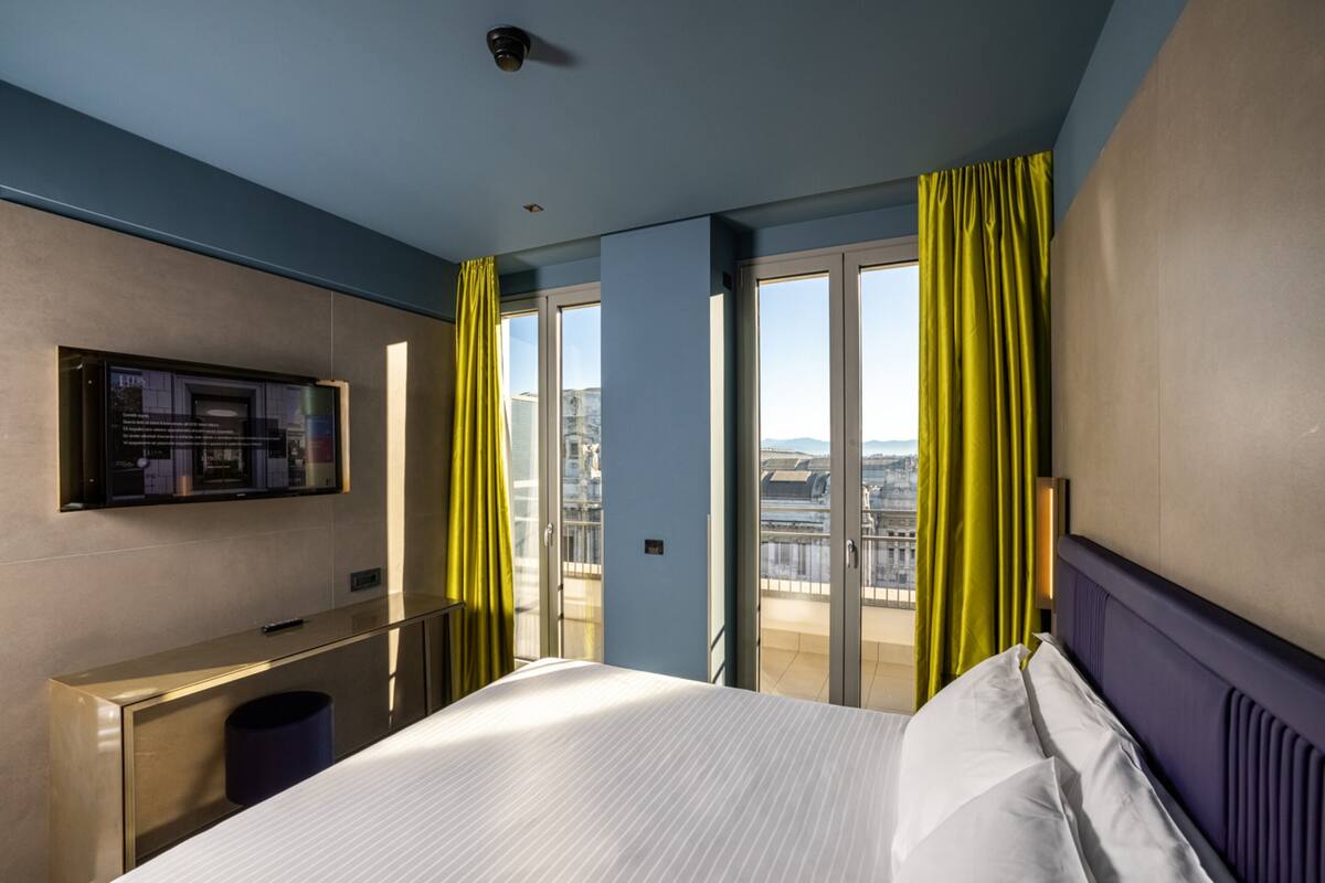 A hotel room with a bed, a desk, a television, and a balcony with curtains.