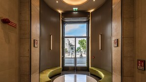 Un hall d’entrée avec une porte en verre, des sièges verts et un reflet sur le sol.