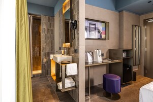 Une salle de bain moderne dans un hôtel, équipée d’une douche, d’un lavabo et de toilettes. On y trouve un meuble-lavabo avec un miroir et un porte-serviettes. Une petite table avec un sac et un téléphone est également visible.