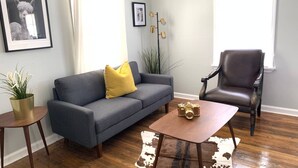 Een woonkamer met een grijze bank, een bruine leren fauteuil, een klein rond tafeltje en een kleden van koeienhuid.