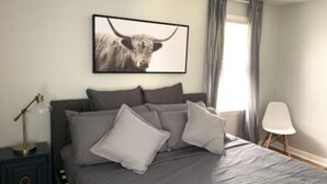 Een slaapkamer met een bed, kussens, een nachtkastje, een lamp, een stoel en een lijst met een afbeelding aan de muur.