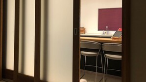 Una habitación con una puerta corredera que da a una zona de cocina con un lavamanos, dos taburetes de bar y una ventana con persianas.