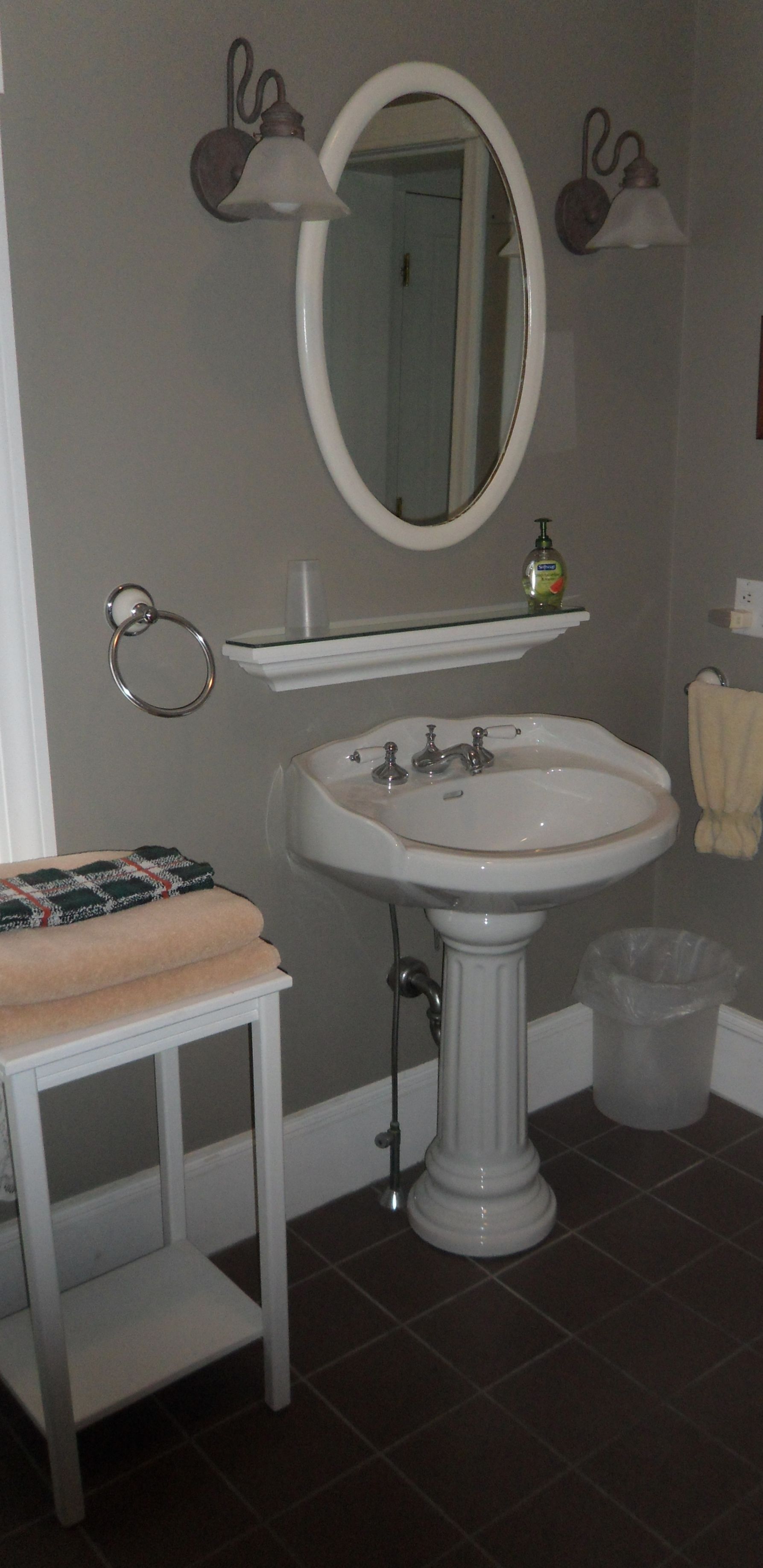 A bathroom with a pedestal sink, a mirror, a towel rack, a glass shelf with a bottle, and a trash bin.