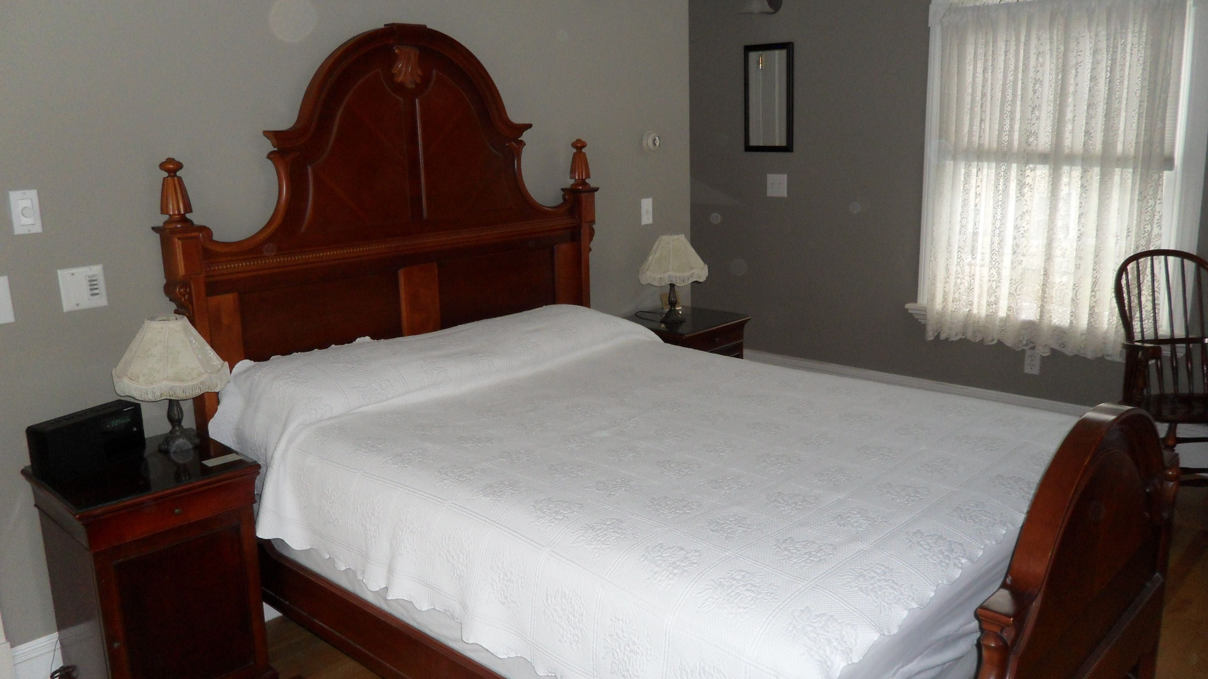 A bedroom with a wooden bed, two bedside tables, a lamp on each, a chair, and a window with curtains.