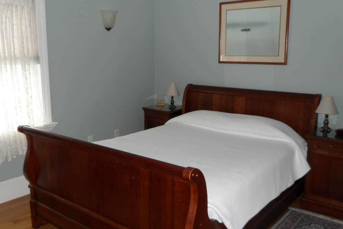 A bedroom with a wooden bed, two bedside tables with lamps, a nightstand, a mirror on the wall, and a window with sheer curtains.
