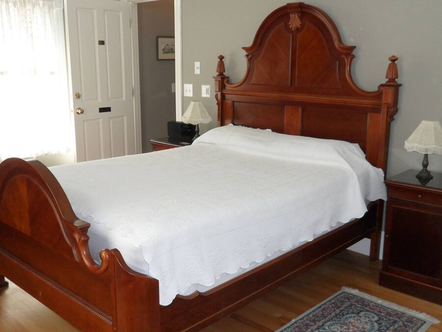 Una cama de madera bien hecha con una colcha blanca, una mesita de noche con una lámpara y una pequeña cómoda.