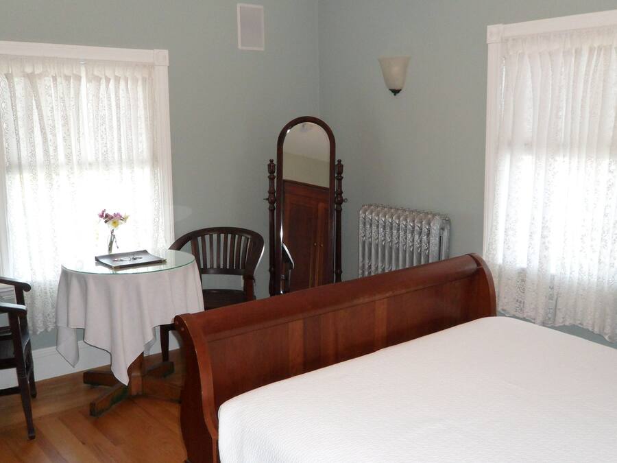 Ein Schlafzimmer mit einem hölzernen Bett, einem runden Tisch mit Stuhl, einem Spiegel und zwei Fenstern mit durchsichtigen Vorhängen.
