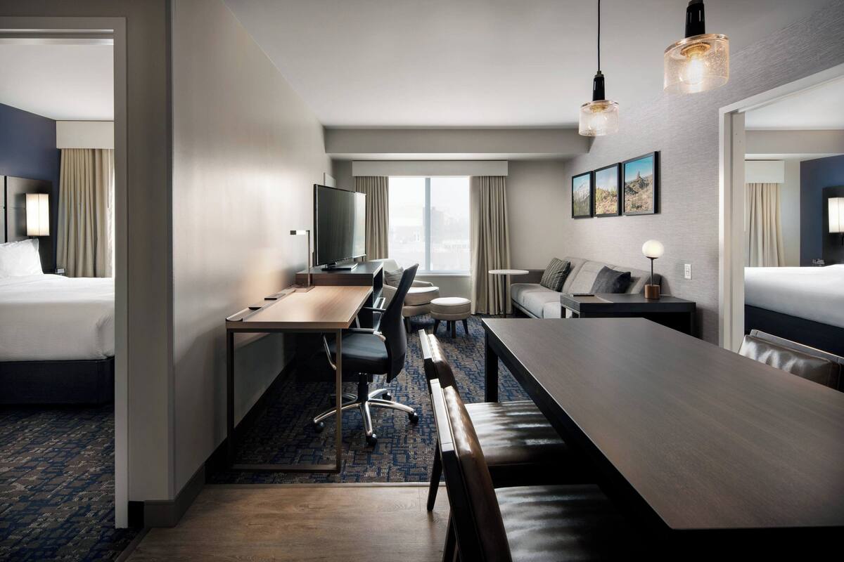 A hotel room with a large bed, a desk with a chair, a dining table with benches, a TV, and a view of the city through the window.
