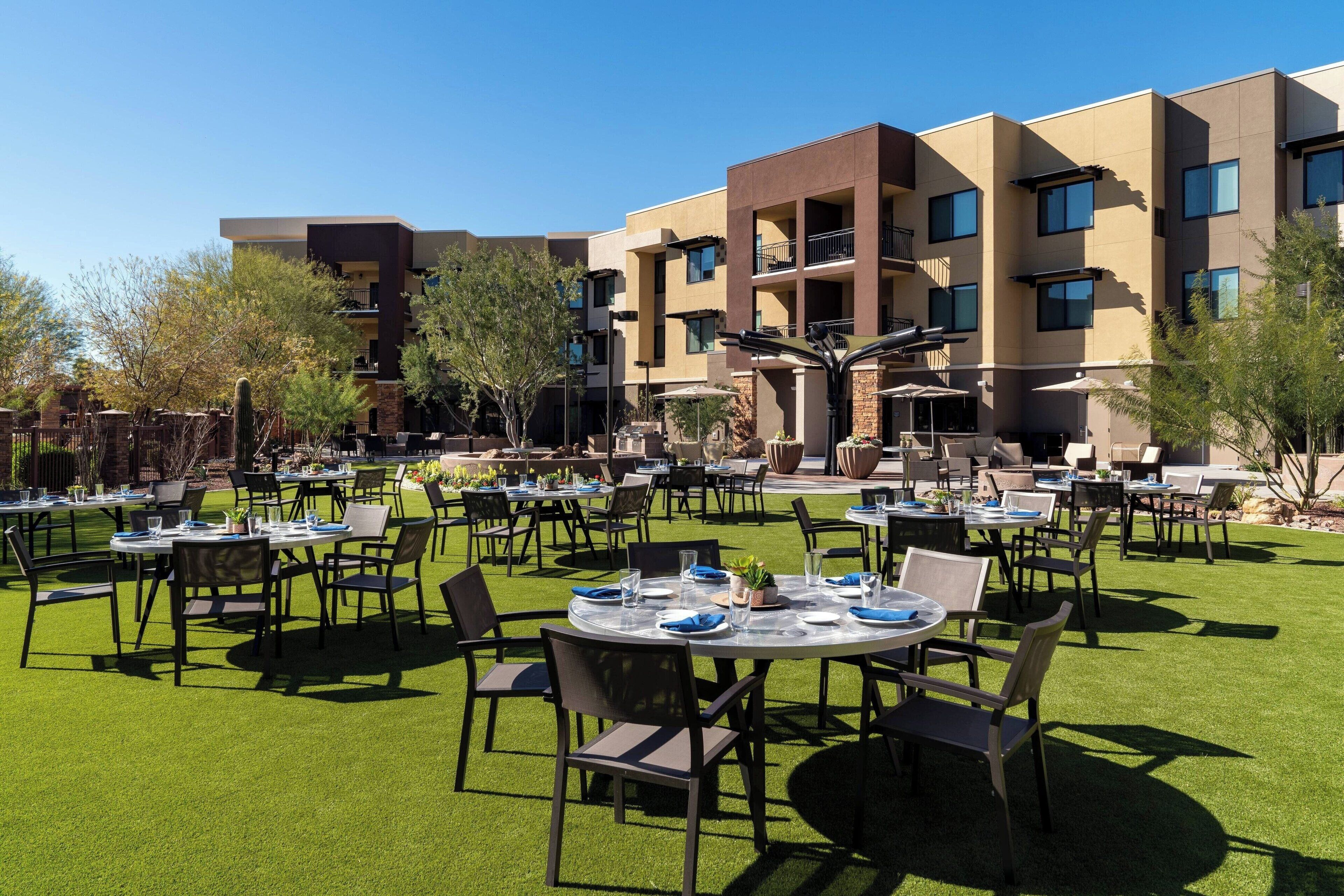An outdoor dining area with round tables and chairs, surrounded by a well-maintained lawn and a multi-story building in the background.