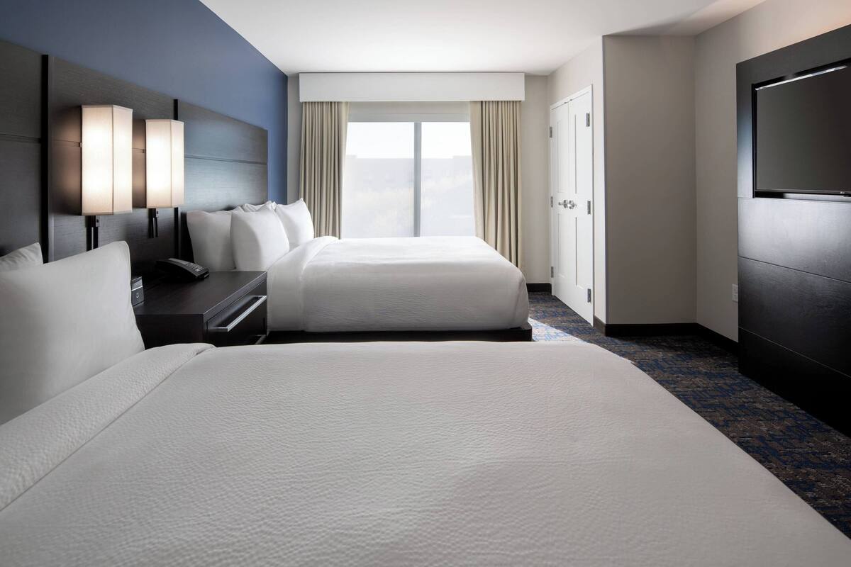 A hotel room with two beds, a desk with a phone, a TV, and a large window with curtains.