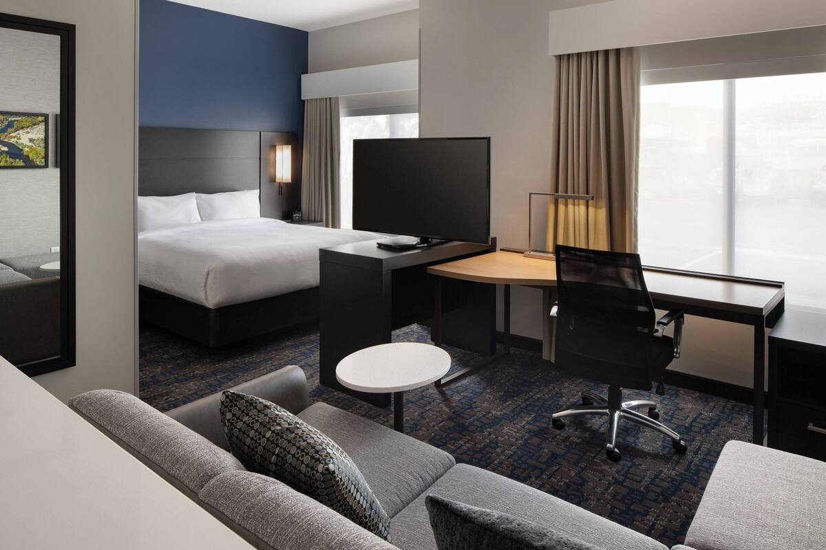 A hotel room with a bed, a desk with a chair, a TV, a sofa, and a small table.