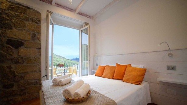 A bedroom with a large bed, a stone wall, a balcony with a table and chairs, and a view of the landscape. - B&B La Roccia dell'Elefante (Castelsardo)