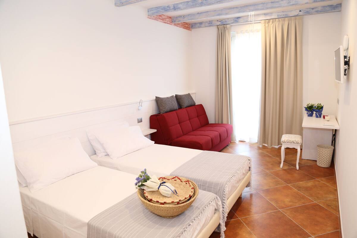A hotel room with two beds, a red sofa, a small table, and a window with curtains.