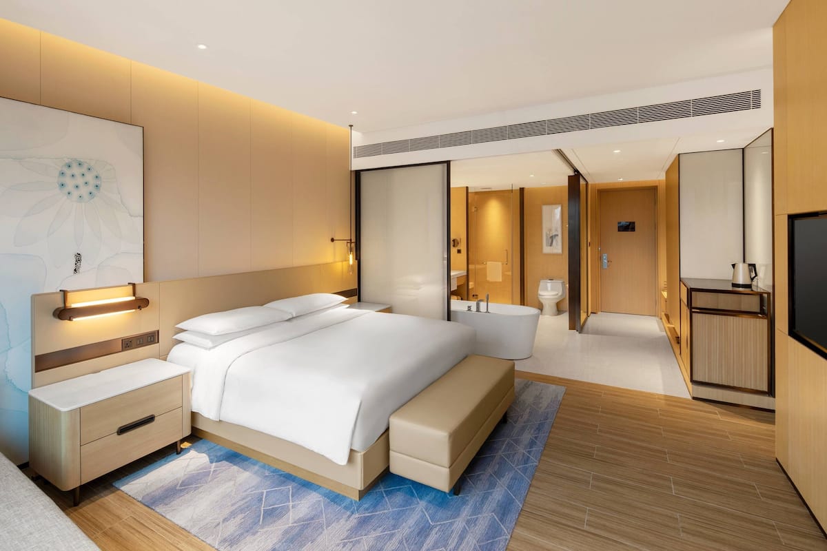 A modern hotel room with a large bed, bedside tables, a built-in bench, a bathroom with a bathtub, and a flat-screen TV.
