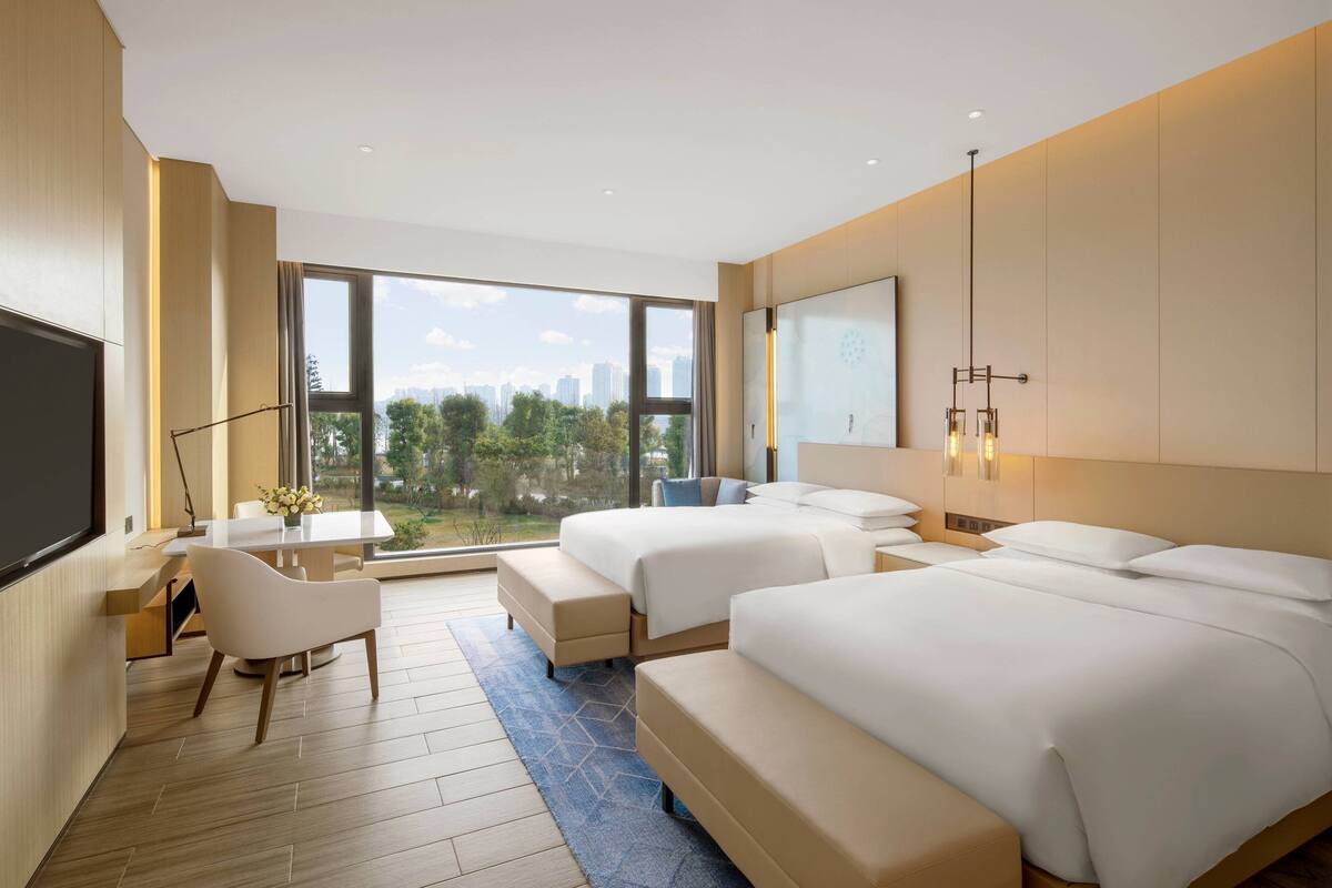 A modern hotel room with two beds, a desk, and a large window offering a city view.