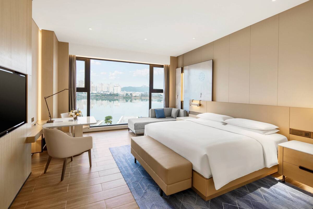 A modern hotel room with a large bed, a desk, a chair, and a view of a body of water.