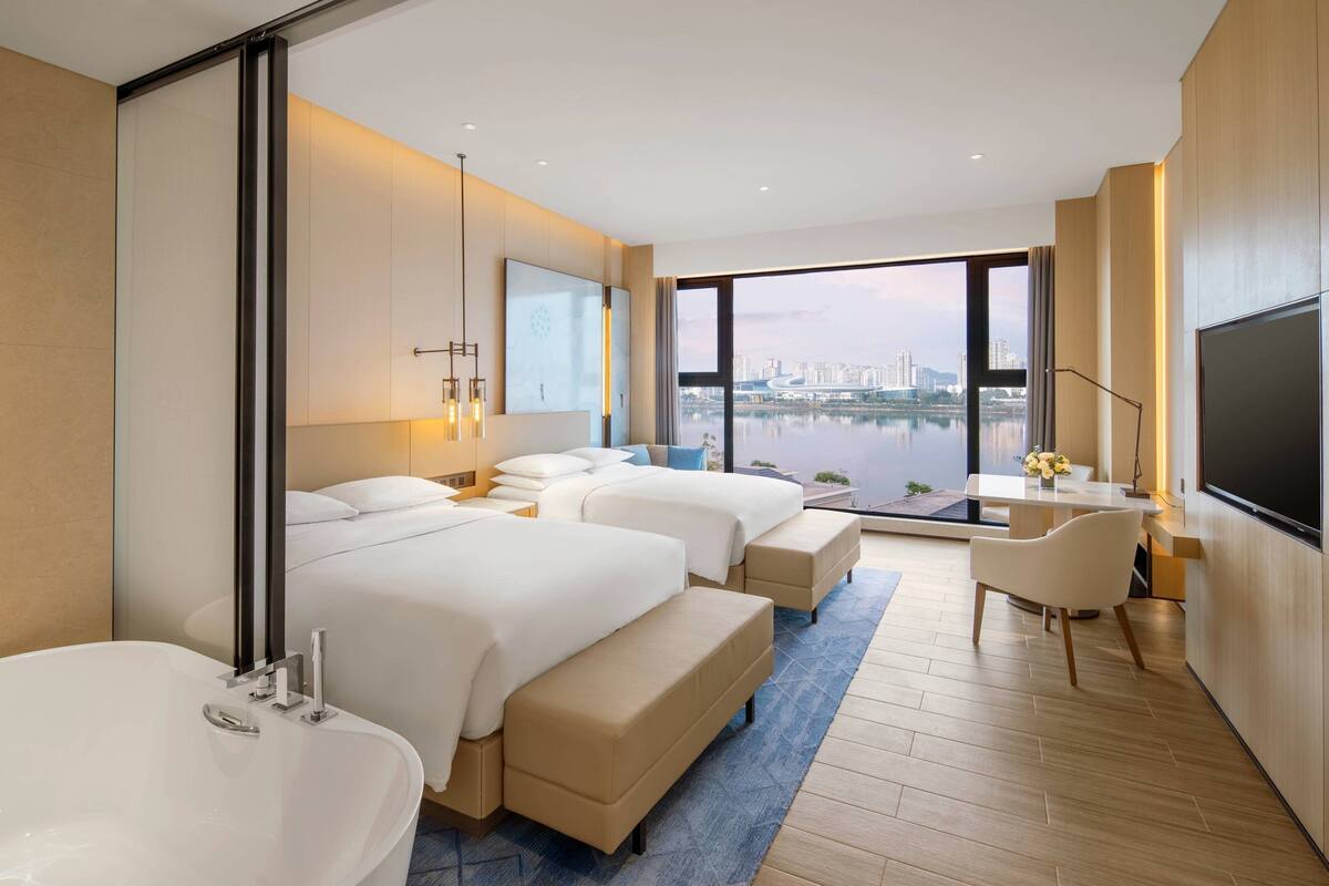 A modern hotel room with a large bed, a separate sofa bed, a desk, and a TV.
