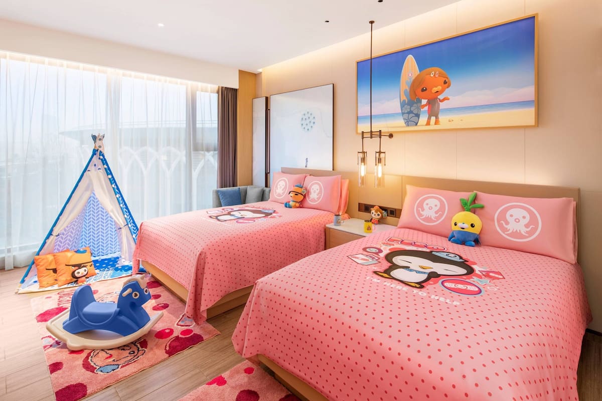 A hotel room with two beds, each with pink bedding featuring cartoon characters. There is a tent set up in the corner, a rocking horse, and a large window with sheer curtains.