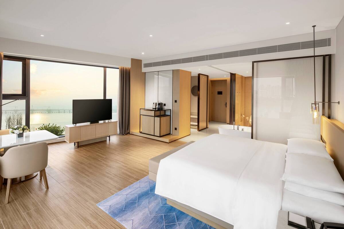 A modern hotel room with a large bed, a desk with a TV, and a view of a bridge.