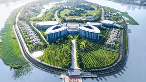Luchtbeeld van een pand aan het water met een centraal, rond gebouw, omgeven door weelderige begroeiing en een bochtige weg.