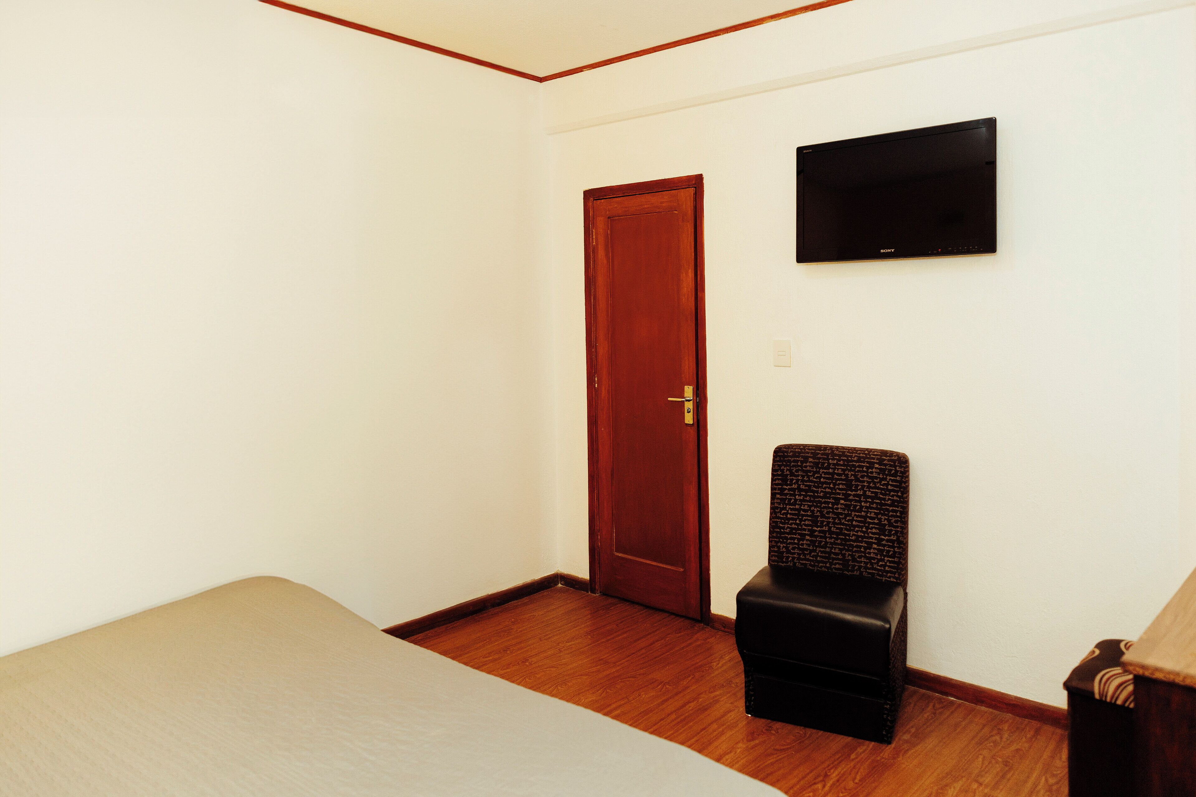 Habitación con suelo de madera, una cama, una silla, una puerta de madera y un televisor de pantalla plana montado en la pared.
