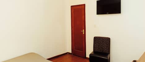 A room with a wooden floor, a bed, a chair, a wooden door, and a mounted flat-screen TV.
