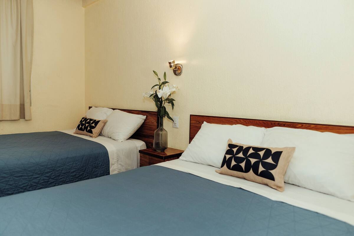Quarto de hotel com duas camas, cada uma com colcha azul e almofadas brancas, mesa de cabeceira com um vaso de flores e luminária fixada na parede.