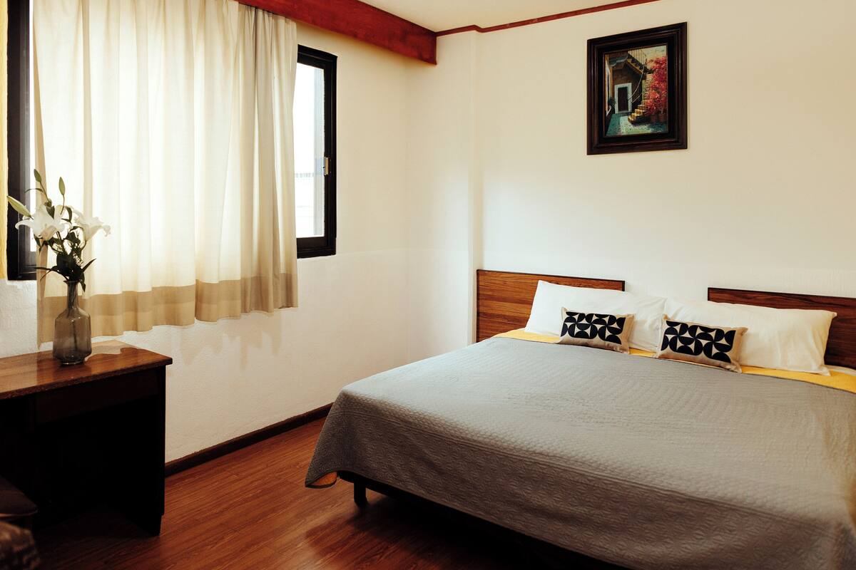Um quarto bem arrumado com uma cama, mesa de cabeceira, janela com cortinas e um quadro na parede.
